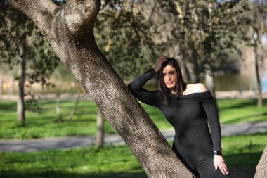 Siyah tulumlu genç, güzel bir kadın, rahat ve sakin, bir ağacın gövdesine yaslanmış poz veriyor. Güzelliği, modayı, barışı, rahatlığı, huzuru kavra.