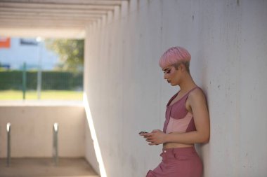 Genç, çekici, eşcinsel, makyajlı, pembe saçlı, üst ve kot pantolonlu, cep telefonunu kontrol eden, duvara yaslanan. LGTBIQ + konsepti, gay, gurur, makyaj, moda, trend.
