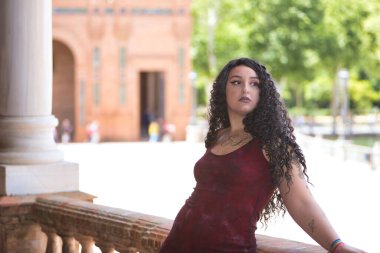 Genç, güzel, alternatif, tombul, kırmızı elbiseli, tuğla korkuluklara yaslanmış, dikkati dağılmış görünen bir kadın. Milenyum konsepti, tombul, bağımsız, farklı.