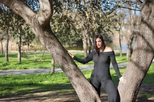 stock image Young, beautiful woman in black jumpsuit, relaxed and calm, posing leaning against the trunk of a tree. Concept beauty, fashion, peace, relaxation, tranquility.