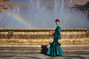 Genç, güzel, tipik yeşil renkli flamenko takım elbiseli sarışın kadın, suya yansıyan gökkuşağı ile bir çeşmenin yanında poz veriyor. Flamenko konsepti, tipik İspanyolca, Sevilla, Endülüs.