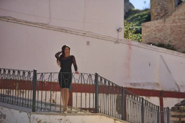 Young, beautiful, brunette woman in an elegant black dress, touching her hair, looking at the camera leaning on a railing in a beautiful white Andalusian village. Concept beauty, fashion, trend.
