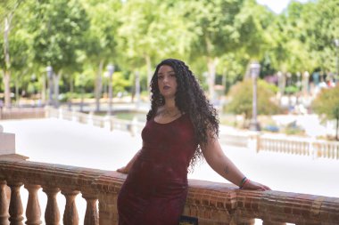 Genç, güzel, alternatif, tombul, dövmeli, kırmızı elbiseli bir kadın, tuğla parmaklıklara yaslanmış, kameraya bakıyor. Milenyum konsepti, tombul, bağımsız, farklı.