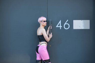 Attractive young gay man, heavily makeup, with pink hair, sunglasses, leather top and pants, posing next to a metal door with number forty-six. LGTBIQ+ concept, gay, pride, makeup, fashion. clipart