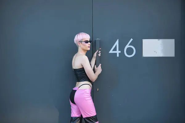 stock image Attractive young gay man, heavily makeup, with pink hair, sunglasses, leather top and pants, posing next to a metal door with number forty-six. LGTBIQ+ concept, gay, pride, makeup, fashion.