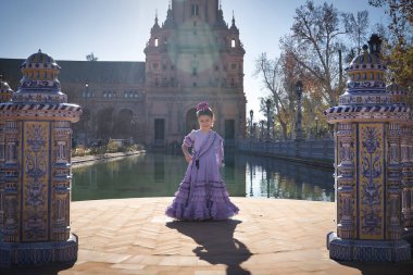 Flamenko yapan kız, kameraya bakıyor, tipik flamenko elbisesiyle, Sevilla 'da güzel bir meydanda, bir gölün yanında güneş ışınları alıyor. Konsept dansı, flamenko, tipik İspanyol dansı..