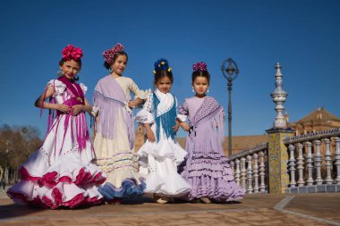 Flamenko yapan dört kız, kameraya bakarak poz veriyorlar, Sevilla 'daki güzel bir meydanda, köprüde tipik flamenko kostümler giyiyorlar. Dans konsepti, flamenko, tipik İspanyolca, Sevilla, İspanya.