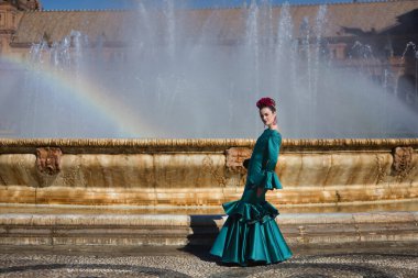 Genç, güzel, tipik yeşil renkli flamenko takım elbiseli sarışın kadın, suya yansıyan gökkuşağı ile bir çeşmenin yanında poz veriyor. Flamenko konsepti, tipik İspanyolca, Sevilla, Endülüs.