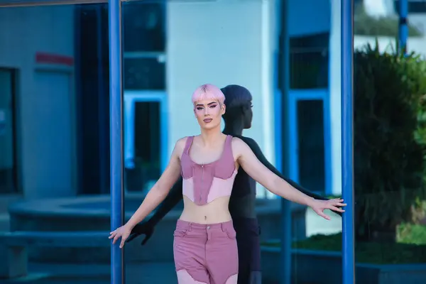stock image Young, attractive, gay man, heavily makeup, with pink hair, top and jeans, looking at camera, leaning against a glass wall with open arms in which he is reflected. Concept lgtbiq+, gay, pride, makeup.
