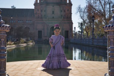 Flamenko yapan kız, kameraya bakıyor, tipik flamenko elbisesiyle, Sevilla 'da güzel bir meydanda, bir gölün yanında güneş ışınları alıyor. Konsept dansı, flamenko, tipik İspanyol dansı..