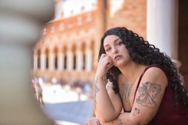 Genç, güzel, alternatif, şişman, dövmeli, kırmızı elbiseli, eli çenesinde tuğla parmaklıklara yaslanmış ve kameraya nazikçe bakan bir kadın. Milenyum konsepti, tombul.