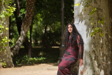 Genç, güzel, alternatif ve tombul bir kadın, kırmızı bir elbise giyiyor, kendini seviyor ve doğayla bağ kuruyor büyük bir ağacın gövdesine yaslanıyor. Bin yıllık, tombul, bağımsız, farklı.