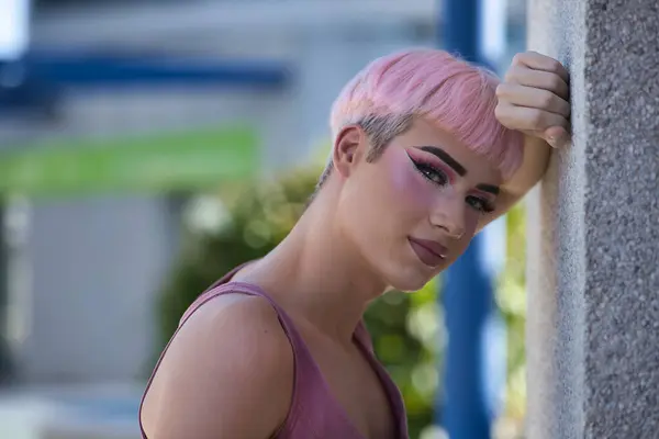 stock image Portrait of young, attractive, gay man, heavily makeup, with pink hair and pink top, leaning against a gray wall looking at camera in close-up, relaxed and calm. Concept lgtbiq+, gay, pride, makeup.