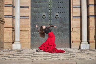 Genç, güzel, esmer, siyah gömlekli, kırmızı etekli, siyah metal bir kapının önünde flamenko dansı yapan bir kadın. Flamenko konsepti, dans, sanat, tipik İspanyolca.