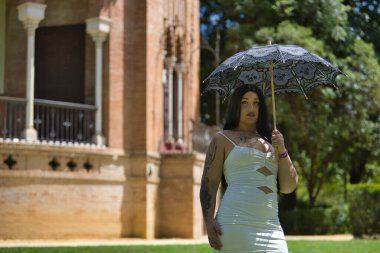 Genç, güzel, alternatif, dövmeli, beyaz elbiseli, antika siyah şemsiyeli, parktaki güneşten korunan tombul bir kadın. Milenyum konsepti, tombul, bağımsız, farklı.