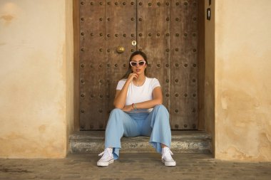 Kahverengi saçlı, güneş gözlüklü, beyaz tişörtlü, mavi pantolonlu, kapı eşiğinde poz veren güzel bir kadın. Güzelliği, modayı, eğilimi kavra.