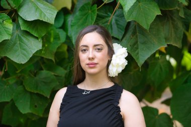 Kahverengi saçlı, siyah takım elbiseli ve yeşil yapraklı arka planda beyaz çiçekli güzel bir kadının portresi. Güzelliği, modayı, trendi, çiçekleri, ilkbaharı kavra.