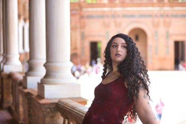 Genç, güzel, alternatif, tombul, kırmızı elbiseli, tuğla korkuluklara yaslanmış, dikkati dağılmış görünen bir kadın. Milenyum konsepti, tombul, bağımsız, farklı.