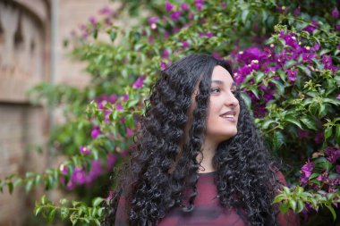 Genç, güzel, alternatif, kırmızı elbiseli tombul bir kadın portresi, dikkati dağılmış görünüyor, bougainvillea arka planında mutlu. Milenyum konsepti, tombul, bağımsız, farklı.