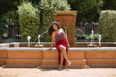 Kadın, genç, güzel, Latin, kıvırcık saçlı, kırmızı elbiseli, mutlu bir şekilde poz veren ve bir çeşmede oturan esmer. Kavram, güzellik, moda, trend.