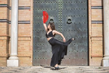 Genç, güzel, esmer, siyah bluzlu ve etekli kadın, eski, siyah metal bir kapının önünde kırmızı pervaneli flamenko dansı yapıyor. Flamenko konsepti, dans, sanat, tipik İspanyolca.