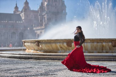 Genç, güzel, esmer, siyah tişörtlü ve kırmızı etekli kadın Sevilla 'daki güzel bir çeşmenin önünde flamenko dansı yapıyor. Flamenko konsepti, dans, sanat, tipik İspanyolca.