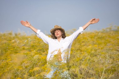 Genç bir kadın, çiftçi, güzel, sarışın, gözleri kapalı, beyaz gömlek, kot pantolon ve şapka giyiyor, açık kollu, özgür ve minnettar, rezene tarlasının ortasında. Güzelliği, minnettarlığı, doğayı kavra..