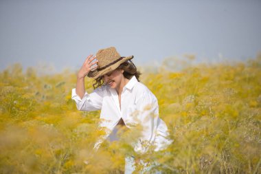 Genç bir kadın, çiftçi, güzel, sarışın, gözleri kapalı, beyaz gömlek, kot pantolon ve şapka giyiyor, rezene tarlasının ortasında mutlu bir şekilde poz veriyor ve gülümsüyor. Güzelliği, mutluluğu, doğayı kavra.