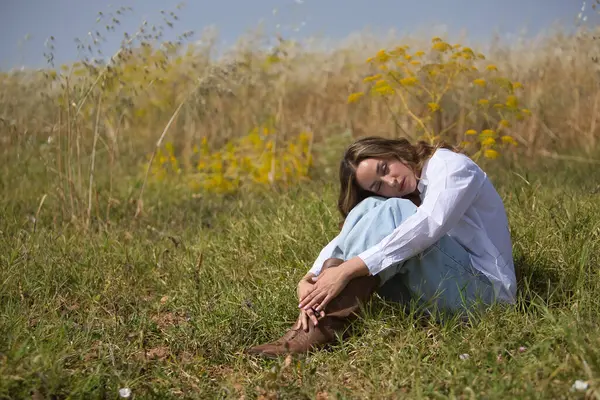 Genç bir kadın, güzel, sarışın, gözleri kapalı, beyaz gömlekli ve kotlu, sahanın ortasında oturmuş, kendine sarılıyor, sakin ve şefkatli. Güzelliği kavra, kırsal, şefkat, huzur, doğa.