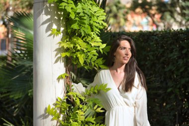 Genç bir kadın, doğal, kahverengi saçlı, beyaz elbiseli, dalgın ve dikkati dağılmış bir vaziyette mermer bir sütuna yaslanmış bitki örtüsüyle. Doğallığı, basitliği, düşünceleri, dikkat dağınıklığını algıla..