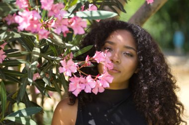 Kadın portresi, genç, güzel, Latin, kıvırcık saçlı esmer, siyah tişörtlü, yanında pembe zakkum. Güzelliği kavra, çiçekleri, ilkbaharı, güzel kokuyu.