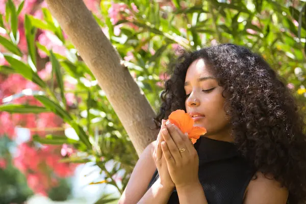 Bir kadın portresi, genç, güzel, Latin, kıvırcık saçlı esmer, siyah bir elbise giyen, bir çiçeğin kokusunu koklayan. Kavram, güzellik, çiçekler, bahar, koku.