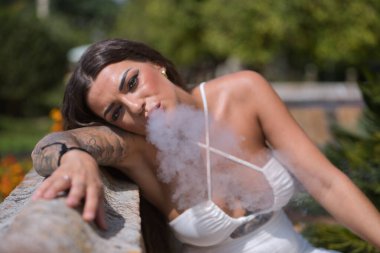 Genç, siyah saçlı, güzel, ağır dövmeli beyaz elbiseli kadın, beyaz buhar dumanı üflerken buhar üflüyor. Dövme konsepti, uyumsuzluk, asilik, trend, moda.