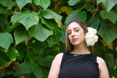 Kahverengi saçlı, siyah takım elbiseli ve yeşil yapraklı arka planda beyaz çiçekli güzel bir kadının portresi. Güzelliği, modayı, trendi, çiçekleri, ilkbaharı kavra.