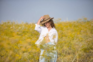 Genç bir kadın, çiftçi, güzel, sarışın, gözleri kapalı, beyaz gömlek, kot pantolon ve şapka giyiyor, rezene tarlasının ortasında mutlu bir şekilde poz veriyor ve gülümsüyor. Güzelliği, mutluluğu, doğayı kavra.