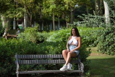 Genç, esmer, güzel, ağır dövmeli, beyaz bluz ve kot giyen, bankta oturan, rahat ve sakin, kameraya bakan bir kadın. Dövme kavramı, uyumsuzluk, isyan, trend.