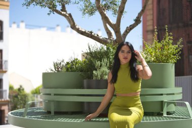 Genç bir kadın, güzel, esmer, latin, yeşil bir takım elbise giyiyor, saçlarına şehvetli bir şekilde dokunuyor, yeşil metal bir bankta bitkilerle oturuyor. Güzelliği, modayı, eğilimi, şehri kavra.