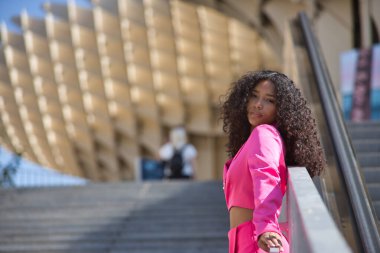 Kadın, genç, güzel, Latin, kıvırcık saçlı, pembe ceketli, rahat ve narin bakışlı, şehirdeki bir merdivende korkuluğa dayanan bir kadın. Güzelliği, modayı, eğilimi kavra.