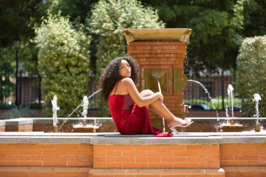 Kadın, genç, güzel, Latin, kıvırcık saçlı, kırmızı elbiseli, çeşmede poz veren. Kavram, güzellik, moda, trend.