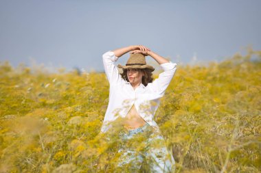 Genç bir kadın, çiftçi, güzel, sarışın, yeşil gözlü, beyaz gömlek, kot pantolon ve şapka giyen, rezene tarlasının ortasında poz veren. Güzelliği kavra, kırsal, doğa.