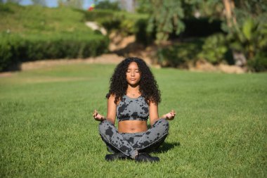 Genç, güzel, esmer, kıvırcık saçlı ve spor giyimli Latin bir kadın, açık hava parkında yoga egzersizleri yapıyor. Konsept spor, rahatlama, meditasyon, egzersiz.