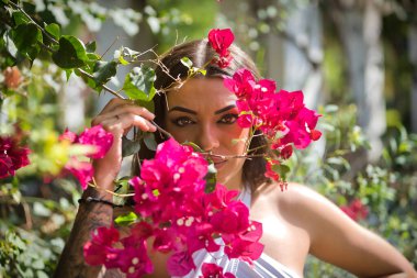 Genç bir kadının portresi, esmer, güzel, çok dövmeli, beyaz bluzlu, begonvillere bakıyor. Kavramsal dövmeler, uyumsuzluk, asilik, trend, çiçekler.