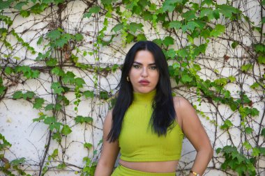 Genç, güzel, esmer, latin kadın portresi, yeşil bir takım elbise giymiş, narin ve narin bakışlı, sarmaşık arkaplanlı bir duvara yaslanmış. Güzelliği, modayı, eğilimi, şehri kavra.