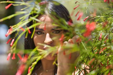 Genç, güzel, esmer, beyaz elbiseli Latin bir kadının portresi, bir bitkinin yapraklarının ve çiçeklerinin arasından bakıyor. Güzelliği, modayı, trendi, çiçekleri, ilkbaharı kavra.