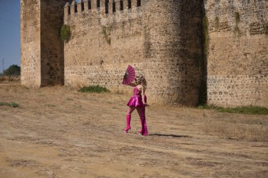 Sürüklenen kraliçe, sarışın, çekici, tacıyla, elbisesiyle, yüksek topuklu botlarıyla ve pembe yelpazesiyle arka planda bir şatoyla dışarı çıkıyor. Gururu, kimliği, çeşitliliği, sanatçıyı kavra.