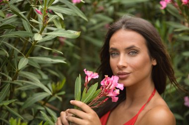 Portrait of a beautiful young woman, with green eyes, in an elegant red dress, smelling the perfume of pink oleanders. Concept beauty, fashion, trend, nature. clipart