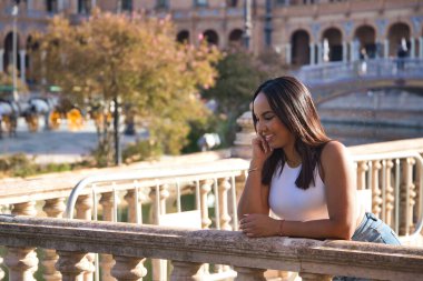 Faslı bir kadın, genç, güzel, esmer, beyaz bluzlu ve kot pantolonlu, eli çenesinde, taş korkuluklara yaslanmış ve mutlu. Etnik köken kavramı, çeşitlilik, kadınlık, güzellik.