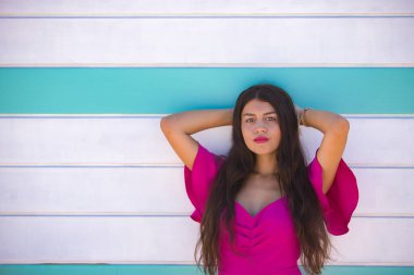 Genç, güzel, esmer, uzun saçlı, pembe takım elbiseli, elleri başının arkasında, arkasında bir plaj kulübesi olan kameraya bakan bir kadın. Güzelliği, modayı, kadınlığı kavra.