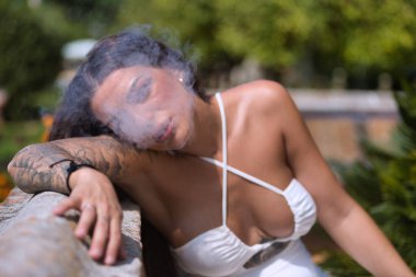 Genç, siyah saçlı, güzel, ağır dövmeli beyaz elbiseli kadın, beyaz buhar dumanı üflerken buhar üflüyor. Dövme konsepti, uyumsuzluk, asilik, trend, moda.