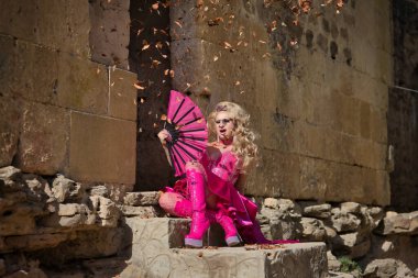 Sürükleme kraliçesi, sarışın, çekici, taçlı, pembe elbiseli ve topuklu ayakkabılı, kuru yaprakların altında büyük bir yelpazeyle kendini yelpazeleyen, taş basamaklarda oturan. Gururu, kimliği, çeşitliliği kavra.